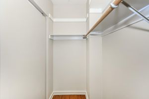 A small, empty closet with light-colored walls, featuring a single wooden clothing rod and a shelf against the back wall. The floor is made of light wooden planks, and the space is well-lit, giving it a clean and organized appearance.