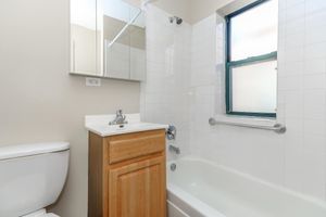 A small bathroom featuring a white bathtub, a wooden cabinet with a sink, and a wall-mounted mirror. The walls are tiled in white, and there is a window with a green frame providing natural light. A grab bar is installed next to the bathtub for safety.