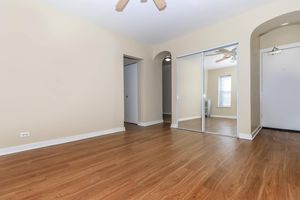 A spacious, empty room with light beige walls and a wooden floor. There are two archways leading to other areas, one with a mirrored closet door. A ceiling fan is visible above. Natural light comes in from a window on the far wall. The door leading outside is on the right.