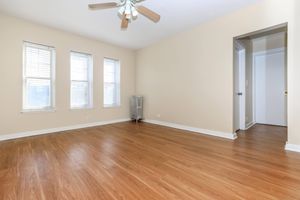 An empty room with light yellow walls and wooden flooring. Three windows with white blinds allow natural light to enter. A ceiling fan is mounted above, and a vintage radiator is visible against one wall. A doorway on the right leads to another space. The overall atmosphere is bright and inviting.