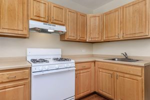 A small kitchen featuring light wood cabinets, a white gas stove, a range hood, and a stainless steel sink. The countertops are beige, and the walls are a soft neutral color, creating a warm and inviting atmosphere.