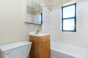 A clean, simple bathroom featuring a white bathtub, a small wooden vanity with a sink, and a mirror above it. There is a toilet beside the vanity, and natural light streams in through a window. The walls are painted a light color, enhancing the bright and airy feel of the space.