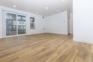 An empty, well-lit living room with large sliding glass doors leading to an outdoor area. The floor is made of light-colored hardwood, and the walls are painted white. There are several recessed lights in the ceiling, and a door leads to another room. The space appears modern and inviting, with ample natural light.