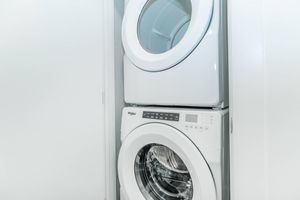 A stacked washer and dryer unit in a modern laundry space, featuring a front-loading design with a sleek, white finish. The washer is on the bottom, while the dryer is positioned above, both enclosed in a light-colored cabinet.