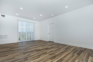 A spacious, empty room featuring large windows with vertical blinds, providing natural light. The flooring is a warm wood finish, and the walls are painted white. A single door leads to another space, and recessed ceiling lights illuminate the area. The overall ambiance is bright and modern.
