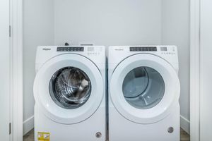 A pair of modern Whirlpool washing machines, one front-loading and one front-loading dryer, neatly positioned side by side in a clean, well-lit laundry room with white walls and gray flooring.