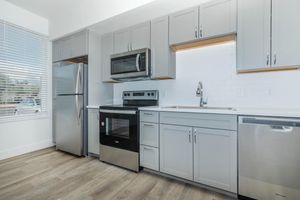 Modern kitchen featuring light gray cabinetry, stainless steel appliances including a refrigerator, microwave, oven, and dishwasher. The countertop is sleek with a white backsplash, and a window allows natural light to brighten the space. The flooring is a warm wood-like laminate.