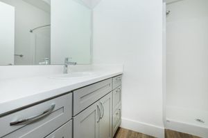 A contemporary bathroom featuring a sleek, white countertop with a sink, silver cabinet drawers, and a large mirror. The design includes a shower area with a glass door and light-colored walls, complemented by modern wood flooring. The overall aesthetic is clean and minimalistic.