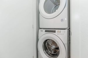 A stackable washer and dryer unit in a modern laundry room, featuring a sleek white design. The washer is positioned at the bottom with a transparent door, while the dryer is above, also with a round glass door. The control panel is visible on the front of the washer, displaying various settings and options.