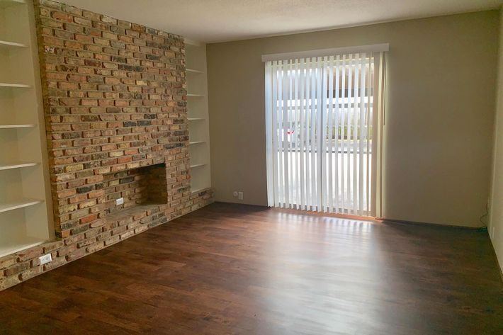 A spacious living room with wooden flooring, a brick wall featuring a small fireplace, and built-in shelves. There are large vertical blinds covering a window that lets in natural light, illuminating the room. The walls are painted in a neutral color, creating a cozy atmosphere.