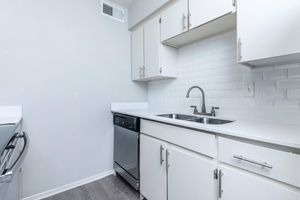 A modern kitchen featuring white cabinetry, a sleek countertop, a double sink with a polished chrome faucet, and a silver dishwasher. The backsplash consists of white tiles, complementing the light color scheme. The flooring is a medium-toned wood, adding warmth to the space.