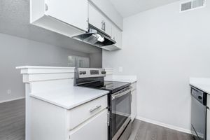 A modern kitchen featuring a stainless steel stove with oven, white cabinetry, and a vent hood. The countertops are light-colored, and the flooring is a light gray laminate. The kitchen is open and connected to a living space with textured walls and a window allowing natural light.
