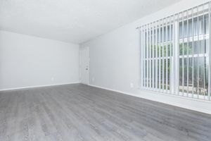 A spacious, empty room with light gray wood flooring, white walls, and vertical blinds covering a large window. The door is visible on one side, and the overall ambiance is bright and minimalistic.