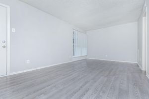 A spacious, empty room with light gray walls and a textured ceiling. The floor features light wood laminate, and there's a large window with vertical blinds. The entry door is visible on the left, and the overall atmosphere is bright and uncluttered, ideal for customization.