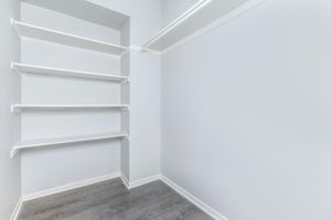 A small, empty closet with light gray walls and white shelves. The floor is a light wood, and the shelves are neatly arranged along one side, creating a clean and minimalistic look. The overall atmosphere is bright and spacious, suitable for storage or organization.