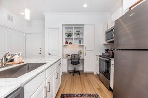 A modern kitchen featuring a spacious countertop with a double sink, stainless steel appliances, and an adjoining workspace. The workspace includes a desk and a black office chair, surrounded by shelves displaying decor and plants. The overall design is bright and inviting, with light wood flooring and minimalistic cabinetry.