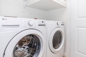 A modern laundry room featuring two white LG washing machines side by side. The machines are stacked under a shelf in a clean, bright space with white walls and doors. One machine is a washer, while the other is a dryer, both equipped with control panels on the front.
