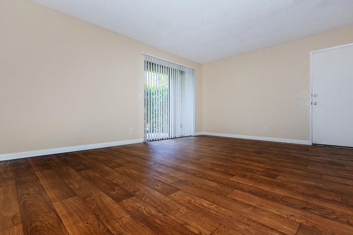 A spacious, empty room with light beige walls and hardwood flooring. There is a large sliding glass door with vertical blinds, leading to a patio area. A white door is visible on the right. The room is well-lit, creating a warm and inviting atmosphere.