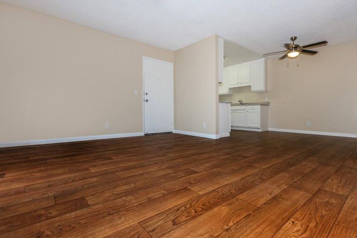 A spacious living area featuring wood laminate flooring, light beige walls, and a ceiling fan. The room has a door leading outside and an open kitchen area visible in the background with white cabinetry. Natural light brightens the space, creating a warm and inviting atmosphere.