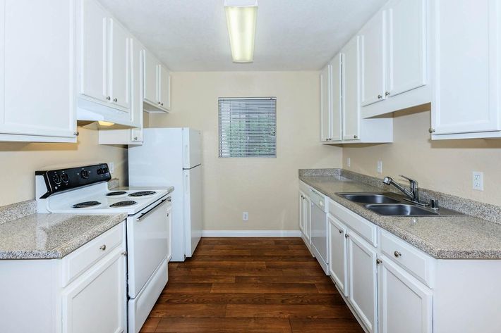 A bright and modern kitchen featuring white cabinetry, granite countertops, a black stove, and a white refrigerator. The kitchen has a double sink and is well-lit with overhead lighting. It includes a window with blinds, and the floor is finished with wood-like planks.