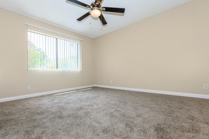 A spacious, empty room with light beige walls and a beige carpeted floor. A ceiling fan with dark blades hangs from the ceiling. A large window covered with vertical blinds allows natural light to fill the space, creating a bright and airy atmosphere. The room has no furniture or decorations.