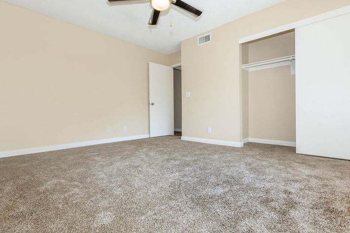 Empty room with beige walls and carpeted floor. A ceiling fan is visible, and there is a white closet on the right. The door on the left is closed, leading to another area. Soft natural light illuminates the space, creating a bright and airy atmosphere.