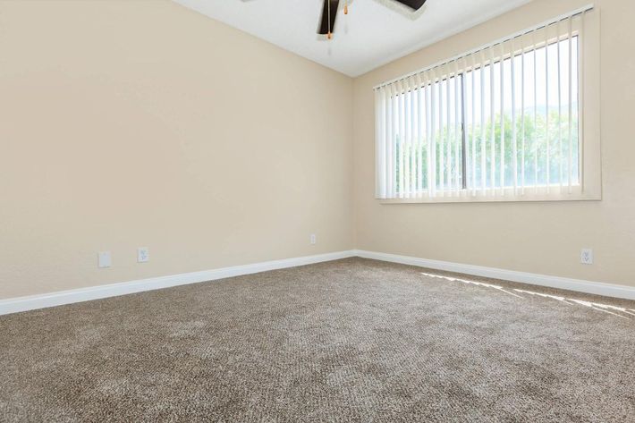 A clean, empty room with beige walls and light brown carpet. A large window with vertical blinds allows natural light to enter, creating a bright atmosphere. The ceiling features a ceiling fan, and there are no furnishings or decorations present, emphasizing the spaciousness of the area.