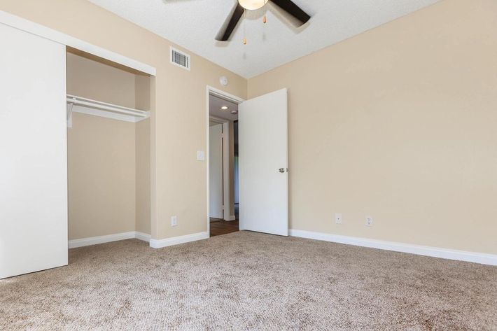 A vacant room with light beige walls and carpet, featuring a ceiling fan. A white door leads to another room, and there's a closet with sliding doors on the left. The space is bright and uncluttered, conveying a feeling of openness and potential for personalization.
