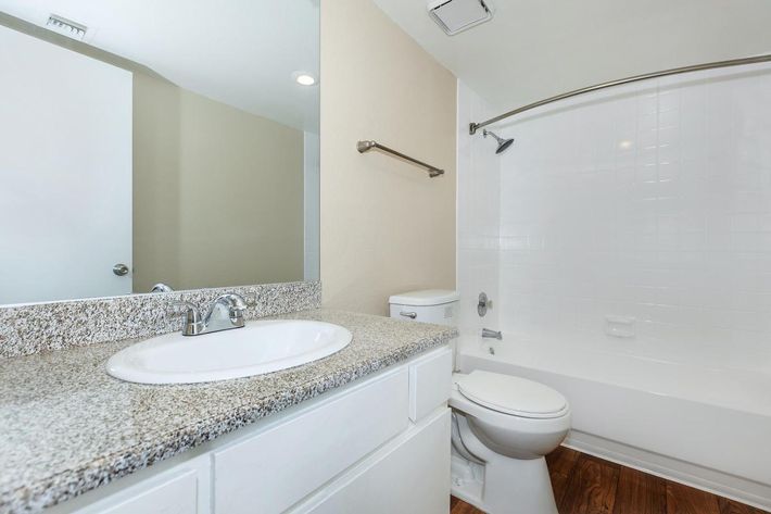 A clean, modern bathroom featuring a white bathtub with a shower, a granite countertop with a sink, and a toilet. Light-colored walls and a mirror reflect a bright, well-lit space, creating an inviting atmosphere. The floor is made of wood, adding warmth to the overall design.
