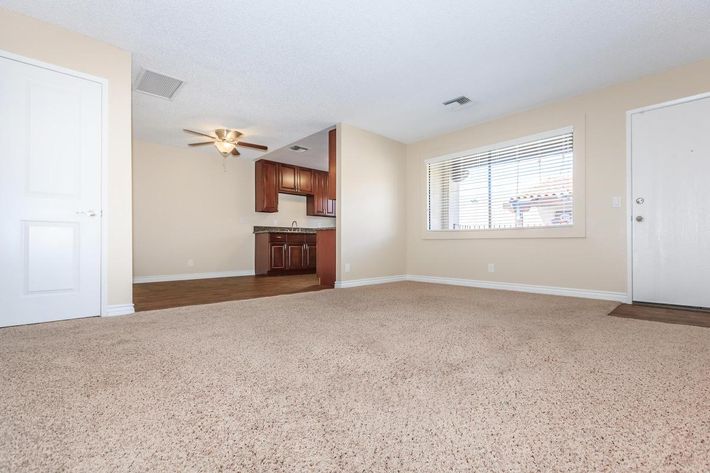Spacious living room with light-colored carpet, featuring a ceiling fan and large window. The background shows an open kitchen with wooden cabinetry. Light-colored walls create a bright, inviting atmosphere. The entrance door is visible on the right.