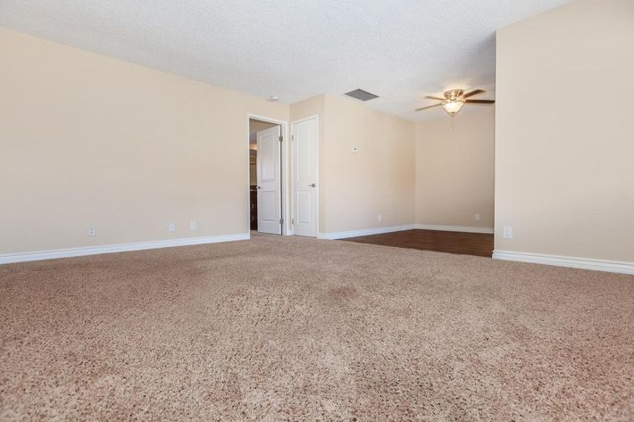 A spacious, empty room with light beige walls and plush carpet. A ceiling fan is visible in the corner, and there are two doorways leading to other areas. Natural light fills the space, creating a warm and inviting atmosphere. The room features a simple and clean layout, perfect for various interior designs.