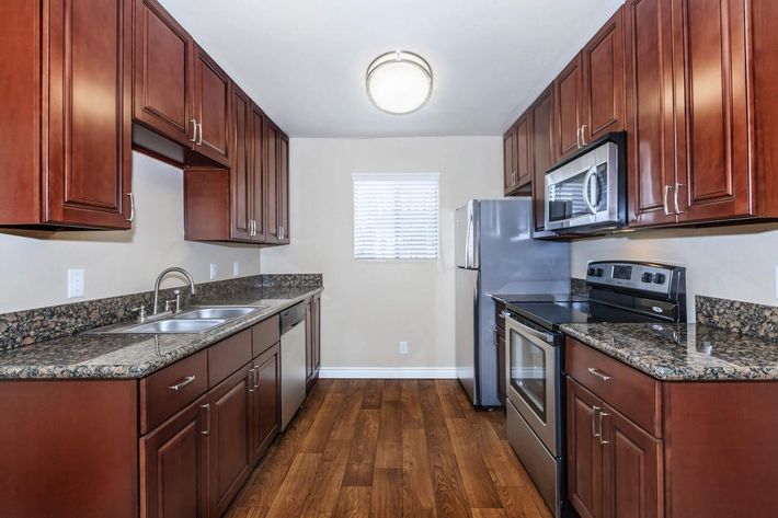 A modern kitchen featuring dark wooden cabinets, granite countertops, stainless steel appliances including a refrigerator, microwave, and oven. The space has a double sink and a window with blinds, illuminated by a ceiling light. The floor is made of wood, enhancing the contemporary design.