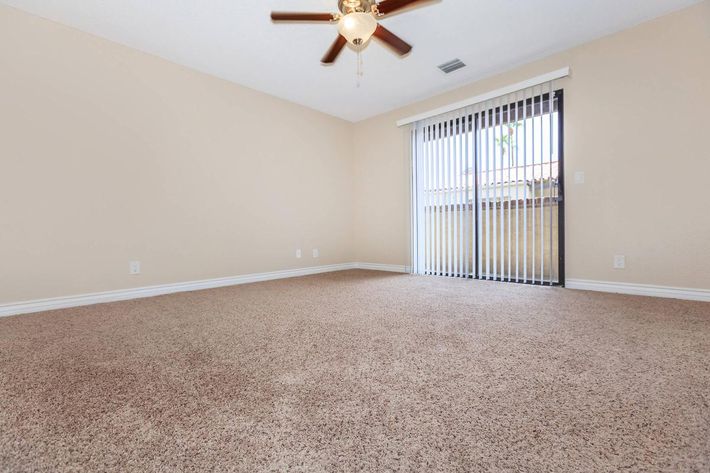 A spacious, empty room with beige walls and light brown carpet. A ceiling fan with five blades hangs from the ceiling. Large sliding glass doors with vertical blinds lead to an outdoor area, allowing natural light to enter the room. The atmosphere feels open and airy, suitable for various uses.