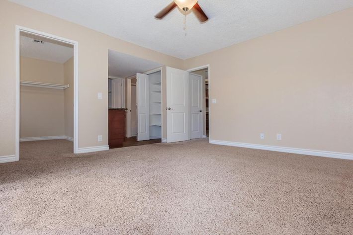 Empty room with beige walls and plush carpeting. A ceiling fan is visible, and there are open double doors leading to another space. To the left, there is an empty closet area. Natural light enters the room, creating a bright but simple atmosphere, ideal for customization or decoration.