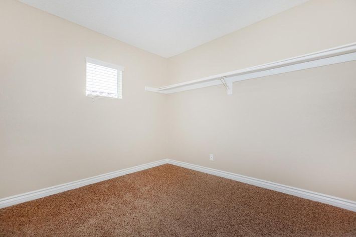 A small, empty room featuring beige walls and a window with white blinds. The floor is covered in light brown carpet, and there's a simple white shelf installed against one wall, providing storage options. The overall atmosphere is clean and minimalistic.