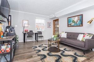 A cozy living room featuring a gray sofa with floral cushions, a round coffee table, and a patterned rug. There's a console table with a coffee machine and decorative items. Two chairs are positioned next to a window, and framed wall art adds color to the space. The room has natural light and a welcoming atmosphere.