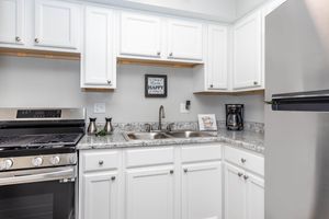 A modern kitchen featuring white cabinetry, granite countertops, a stainless steel refrigerator, a gas stove, and a sink. The kitchen is well-lit, with open shelves above the sink displaying a small decorative sign and a coffee maker.