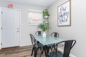 A cozy dining area featuring a glass-top table with a white vase filled with purple flowers, surrounded by four black metal chairs. The wall displays a framed sign with the text "Our happy place." A window with blinds allows natural light, and an exit sign is visible near the door.