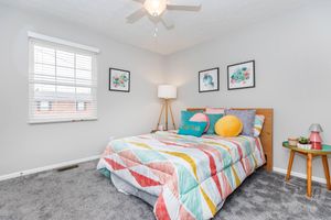 A cozy bedroom featuring a colorful, patterned bedspread with decorative pillows. There's a nightstand with a small lamp and potted plant, along with framed floral art on the walls. A window with blinds allows natural light, and a ceiling fan is installed overhead. The carpet is plush and gray.