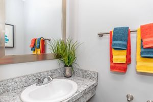 A modern bathroom featuring a granite countertop with a white sink. Two vibrant towels in orange, blue, and yellow hang on a towel rack. A small green plant in a decorative pot adds a touch of greenery, and a mirror reflects the space. The walls are painted a light color, enhancing the bright atmosphere.