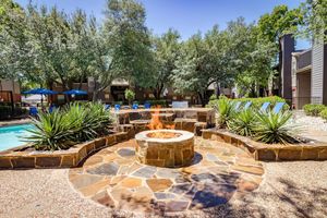 A landscaped outdoor area featuring a circular stone fire pit surrounded by seating, with lush greenery and trees in the background. Blue lounge chairs and umbrellas are visible near a swimming pool, creating a relaxing atmosphere.
