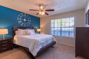 A well-decorated bedroom featuring a large bed with white bedding, two bedside lamps on dark wooden nightstands, a blue accent wall with a large clock, and a window allowing natural light to enter. The room has carpeted flooring and a wooden dresser.