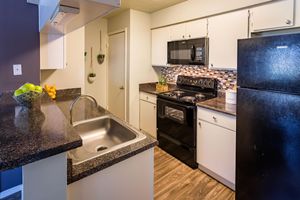 A modern kitchen featuring a black refrigerator and stove, a stainless steel sink, and dark countertops. The cabinets are white with a backsplash of colorful tiles. A bowl of green apples and a cluster of lemons sit in a decorative bowl on the countertop. Natural light enhances the spacious feel of the kitchen.