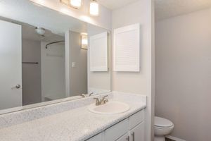 A modern bathroom featuring a white countertop with a sink, two mirrors above it, and light fixtures. The walls are painted in neutral tones, and there is a white toilet visible. A shower area is seen in the background, with a sliding glass door.