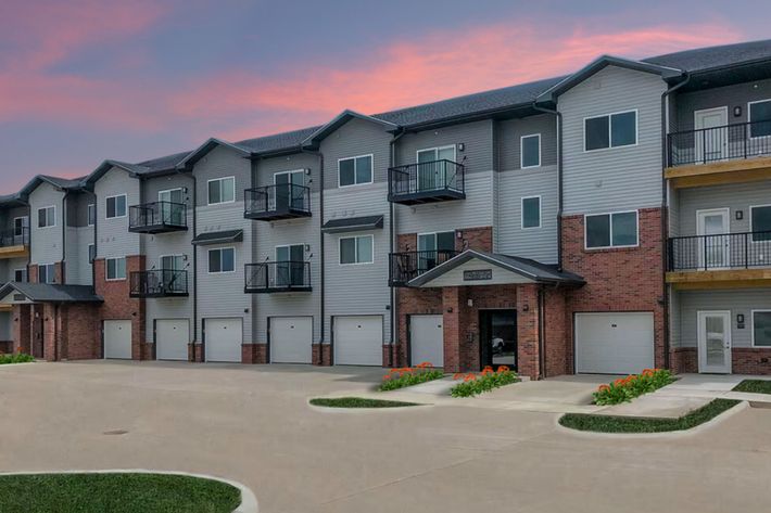 A modern apartment building with multiple units, featuring a mix of brick and siding exteriors. Each unit has a small balcony, and there are garages on the ground level. The background shows a colorful sunset sky. The surrounding area is landscaped with green grass and flowers.