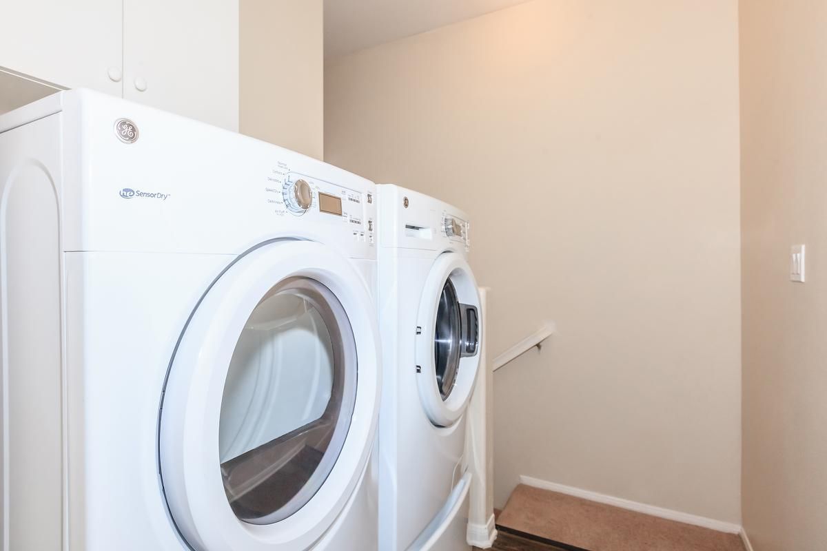 A laundry room featuring two modern white washing machines side by side, against a light beige wall. There’s a staircase visible in the background, and the space appears well-lit and organized.