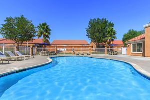 A clear blue swimming pool surrounded by lounge chairs, palm trees, and residential buildings with red tile roofs under a sunny blue sky.