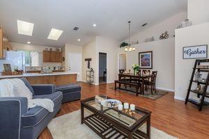 A cozy living room and dining area featuring a blue sofa, wooden dining table with chairs, and a coffee table adorned with decorative items. The space is well-lit with natural light, showcasing an open layout with warm wooden flooring and tasteful decor, including a "Gather" sign on the wall.