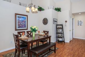 A cozy dining area featuring a wooden table set for two, adorned with a floral centerpiece. A decorative framed picture hangs on the wall, complemented by a clock. The room has light walls, wooden flooring, and greenery accents. A shelving unit with various decor items is positioned nearby.