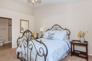 A cozy bedroom featuring a wrought iron bed with white bedding and decorative pillows. There's a small table with a lamp beside the bed, a mirror on the wall, and a vase with flowers. A bathroom entrance can be seen in the background. The walls are painted a warm beige, creating a calm atmosphere.
